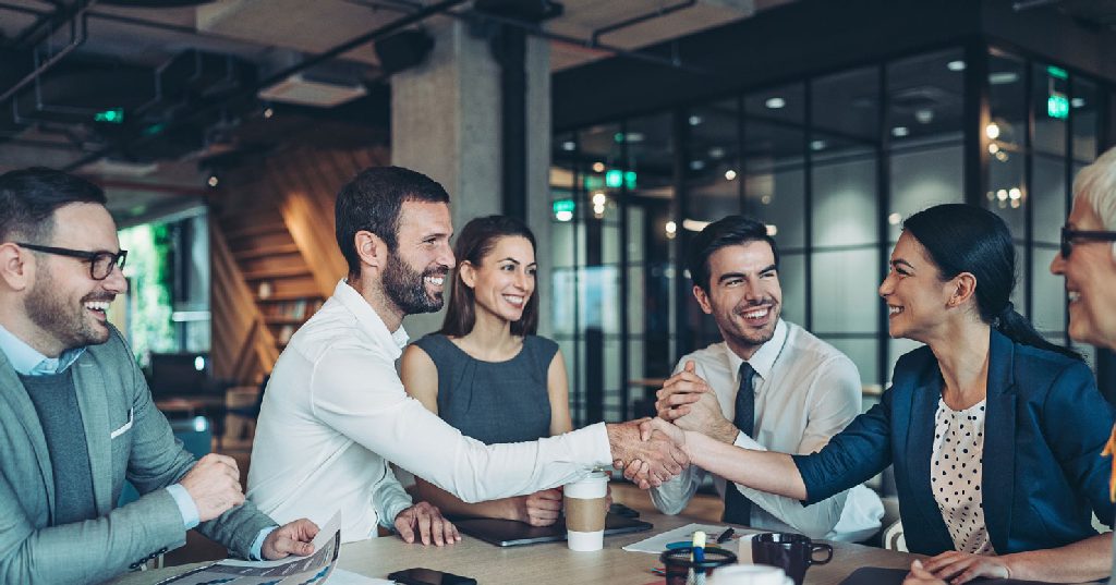 Fünf Geschäftsleute sitzen um einen Tisch in einem Büro mit Glaswänden. Zwei Personen geben sich lächelnd die Hand. Alle haben professionelle Kleidung an und wirken freundlich und engagiert.
